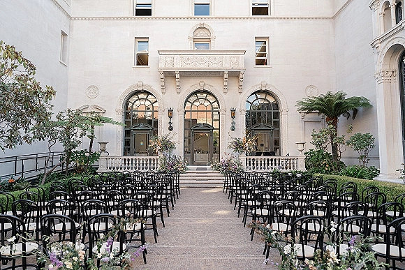 Ceremony setup with black chairs and white cushions lining a floral aisle in a courtyard, facing arched windows and double doors.