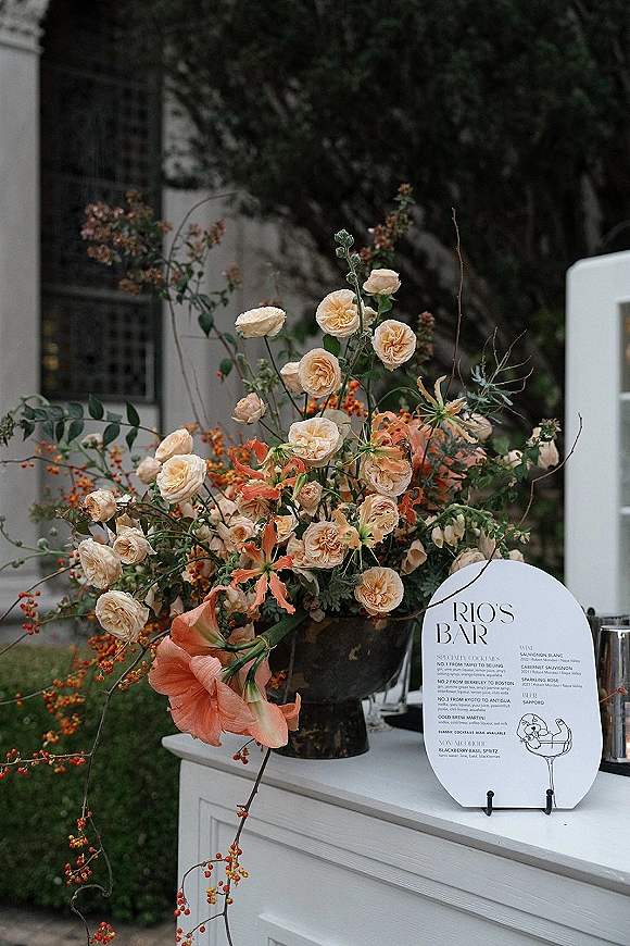 Wedding bar sign with signature cocktail sign details, framed by roses and orange flowers in a vase on the bar counter with candlelight nearby