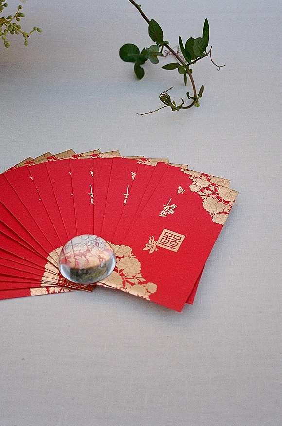 Red envelopes with gold floral print and double happiness symbol arranged on a light gray surface with a glass orb and greenery sprig