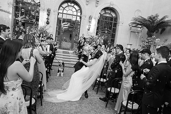 Wedding recessional with bride and groom recessional walking through bubbles, her long veil trailing as guests line courtyard chairs by arched windows