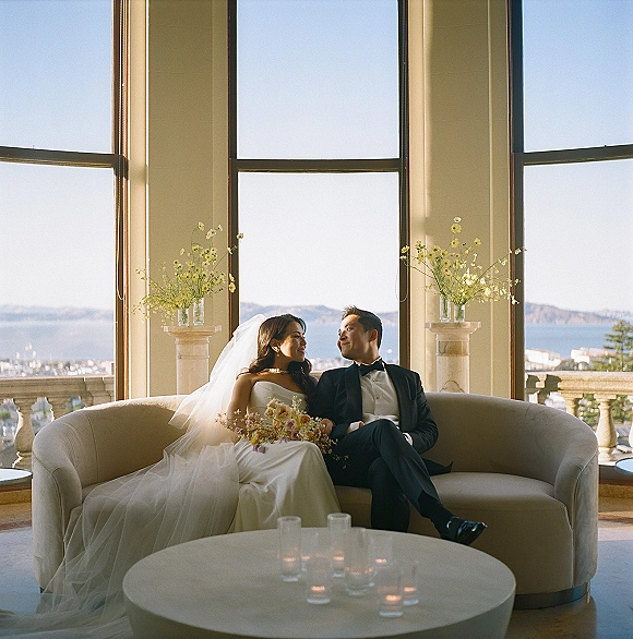 Couple portrait of bride and groom on sofa, her strapless gown and veil with pastel bouquet, by large windows with ocean view beyond