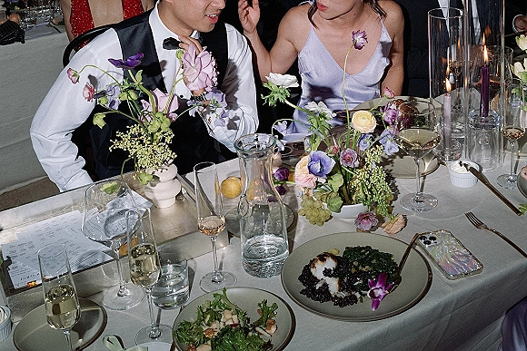 Wedding reception moment as a groom in tuxedo vest chats at a candlelit table with pastel floral centerpiece, menus, and guests on banquette seating