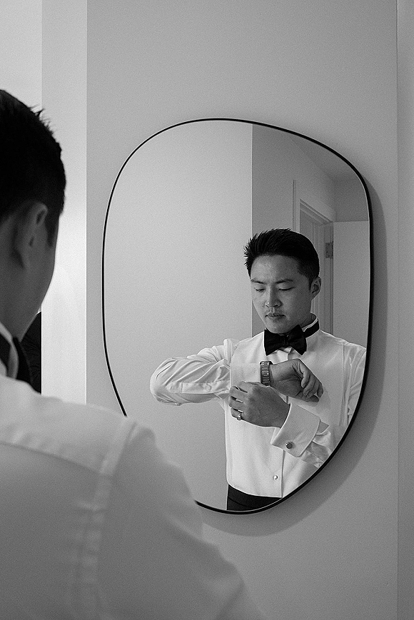 Groom getting ready, groom mirror portrait adjusting cufflinks and checking wristwatch with wedding band in a doorway by a white wall