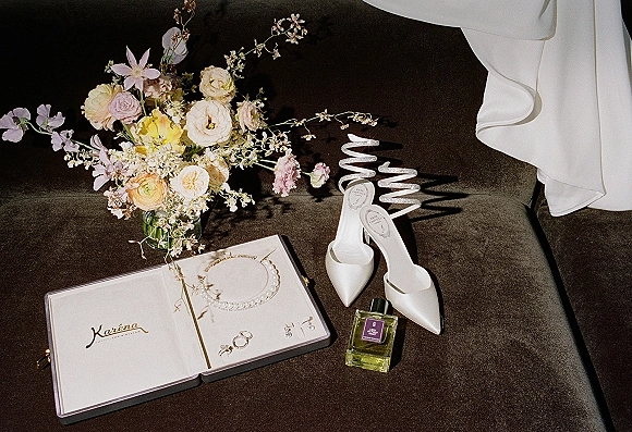 Wedding details flatlay with bridal accessories flatlay, lace-up heels, pearl jewelry, rings, perfume, and pastel bouquet on a dark velvet sofa