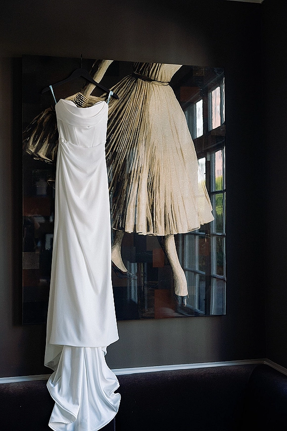 Wedding dress, strapless wedding dress in satin hangs on a dress hanger, showing corset bodice and train against a dark wall with artwork