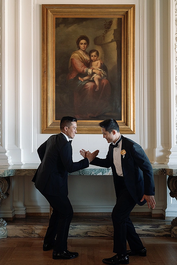 Groom portrait of two grooms shaking hands in black tuxedos and bow ties, boutonniere details by a stone mantel and framed painting