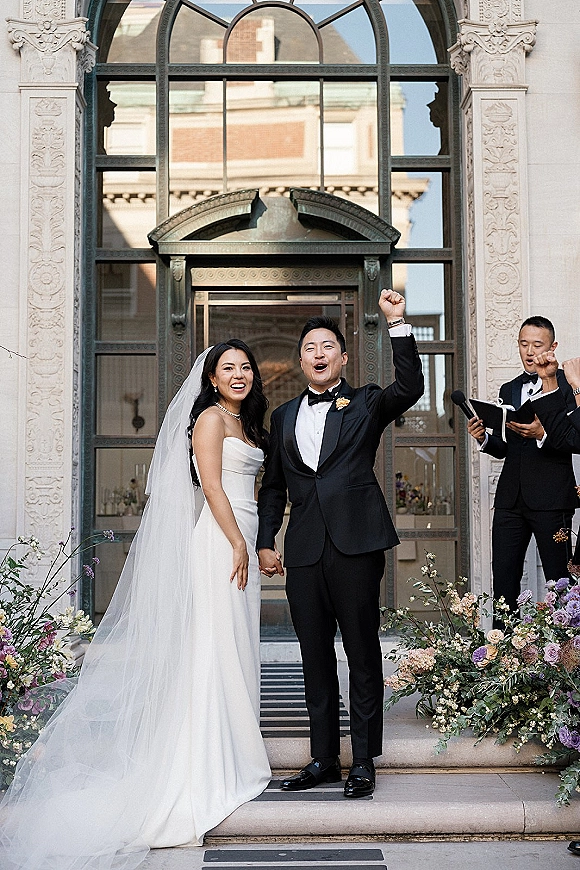 Recessional moment as newlyweds cheer holding hands, bride in veil and groom in tux, stepping through an arched glass doorway on stone steps