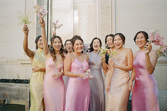 Bridesmaid group photo in pastel bridesmaid dresses, holding small bouquets and laughing by a white facade with tall window greenery