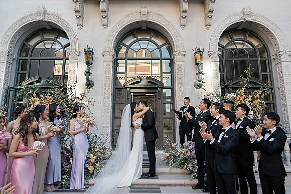 Wedding kiss portrait of bride and groom kissing as bridesmaids in lavender and groomsmen cheer on stone steps, veil flowing behind