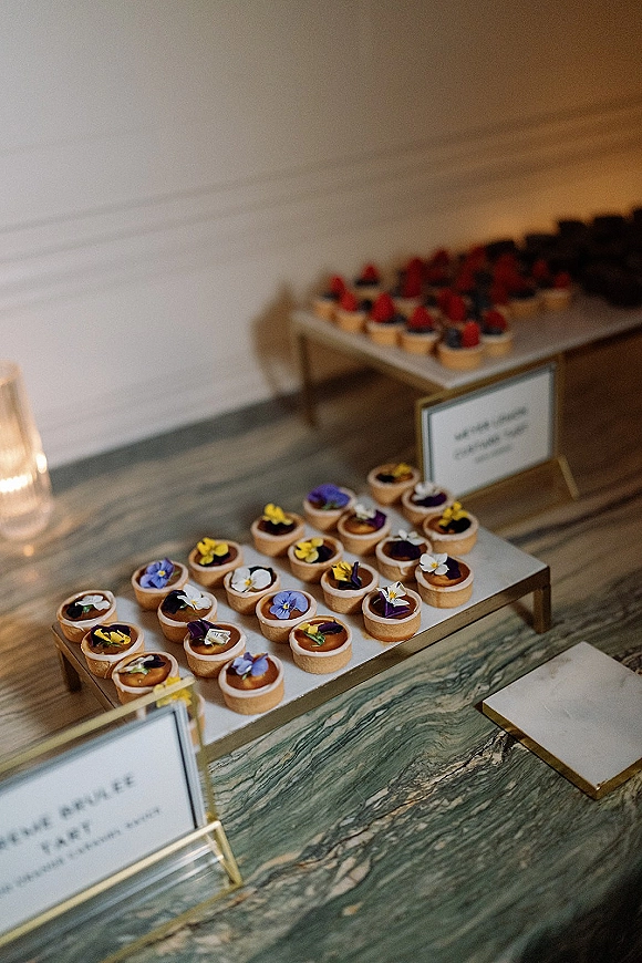 Wedding dessert table with mini tart dessert table trays, berry and edible flower garnishes, gold label frames and a candle votive on marble