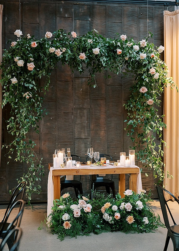 Reception sweetheart table with a candlelit sweetheart table backdrop, blush roses and greenery arch, wooden table set against dark wood paneling