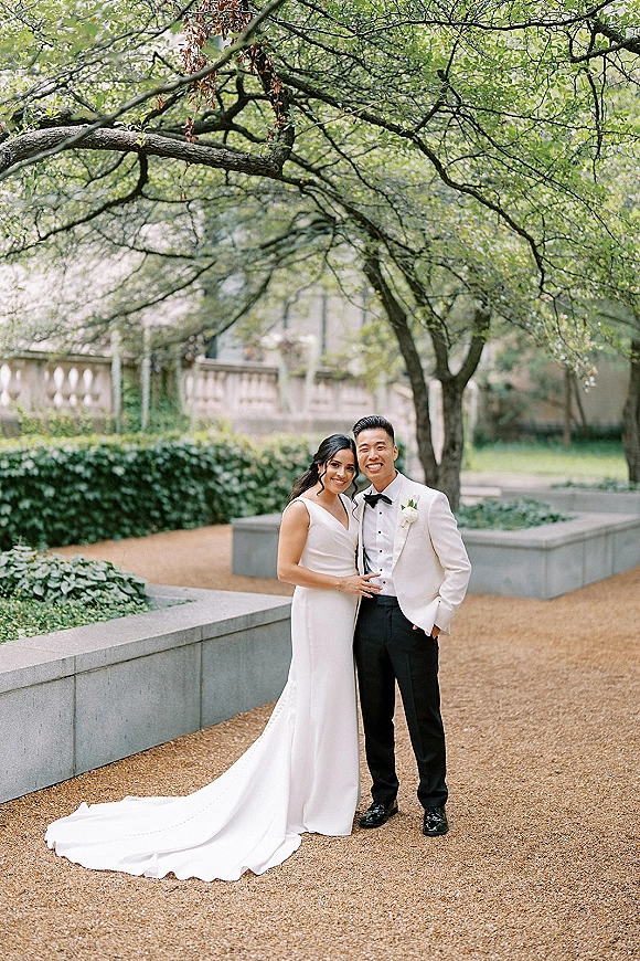 Couple portrait of bride and groom smiling on a gravel garden path, her wedding dress train flowing beside his tuxedo and boutonniere
