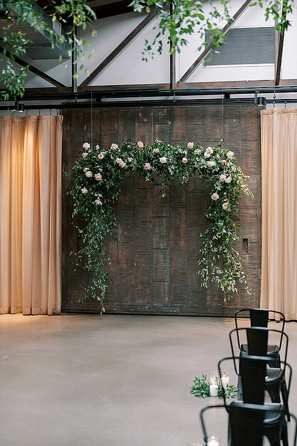 Wedding ceremony backdrop with greenery installation and blush rose garland over draped curtains, candles and chair rows against a wood wall