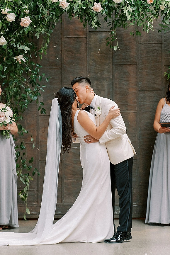 Wedding kiss as the bride and groom share a ceremony kiss moment under a floral arch, her long veil draping before a wood wall backdrop