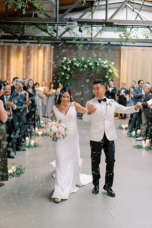 Wedding recessional as bride and groom walk hand in hand through bubbles, passing aisle candles and guests in an industrial indoor venue
