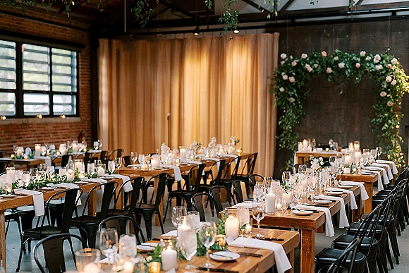 Reception tablescape with wood banquet table decor, white runner, greenery garland and candlelit place settings under string lights by brick wall