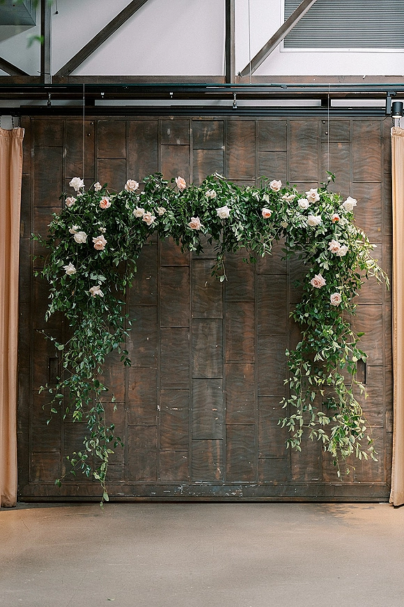Wedding floral arch with greenery wedding arch garlands, blush and white roses, and draped curtains against a dark wood wall backdrop