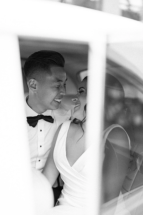 Couple portrait, black and white wedding portrait of bride in sleeveless dress and groom in bow tie laughing inside a car, framed by window
