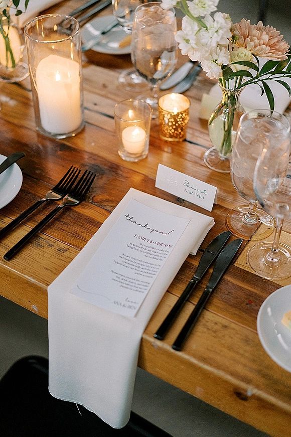Reception tablescape on a rustic wedding table with taper and hurricane candles, floral greenery centerpiece, menu and place cards, and black flatware indoors