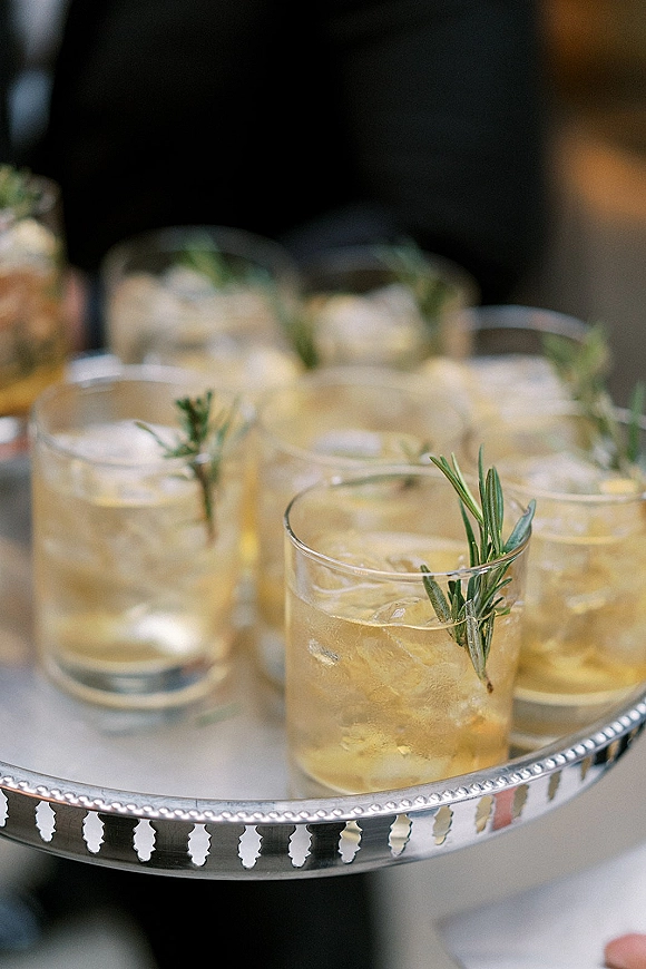 Wedding cocktails served as signature wedding cocktail in lowball glasses with ice and rosemary sprigs on a silver tray in a blurred reception setting