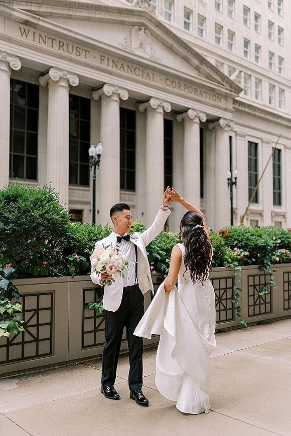 Couple portrait of bride and groom twirl on a city sidewalk by grand columns, bride in strapless gown with blush rose bouquet