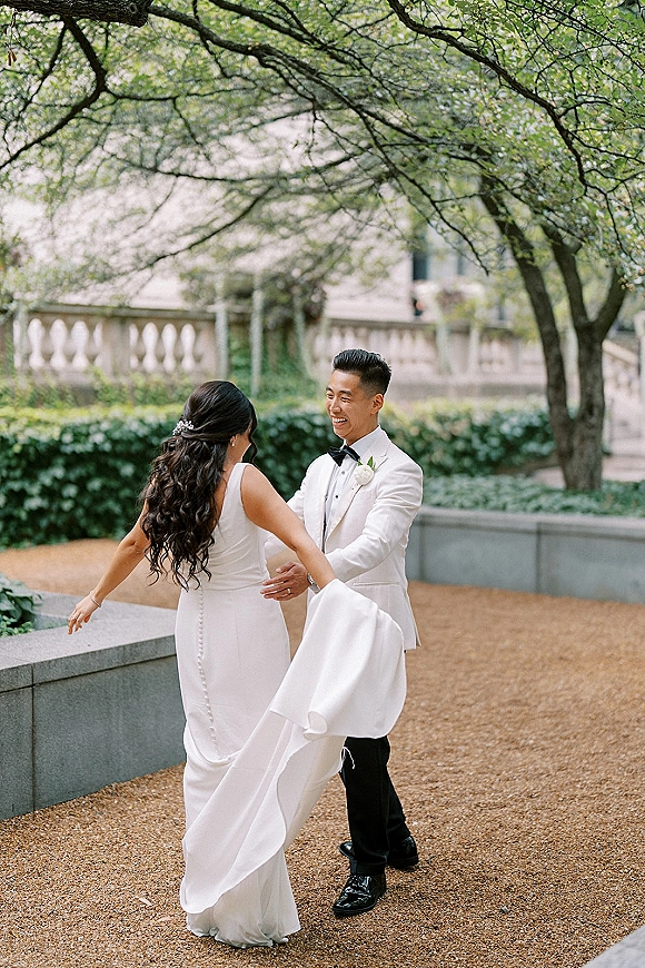 First look moment as bride twirls in a wedding first look, holding her dress train while groom in white tux watches on a garden path