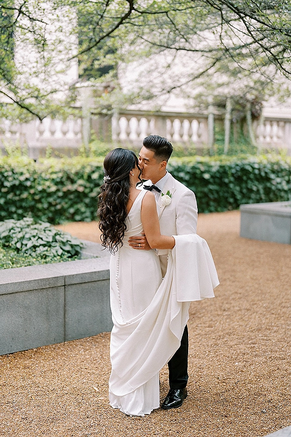 Wedding kiss portrait of bride and groom kissing, her long train draped over gravel path in formal garden with hedges and stone planters