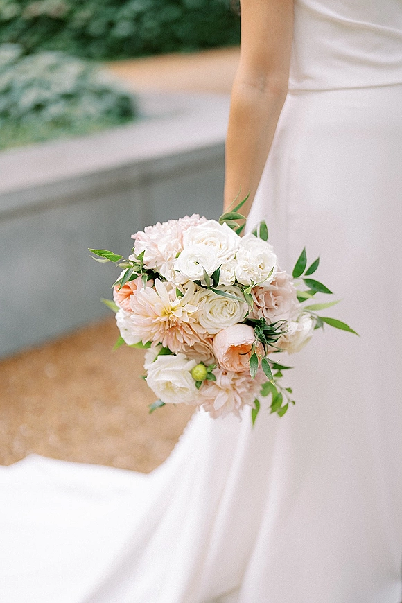 Bridal bouquet of blush and white flowers with greenery held against a white wedding dress on a gravel path by a stone wall
