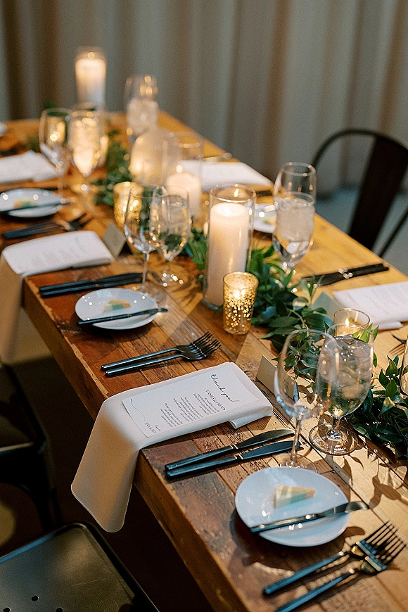 Reception tablescape on a rustic wedding table with greenery garland, pillar and votive candles, menus, place cards, and glassware before curtains