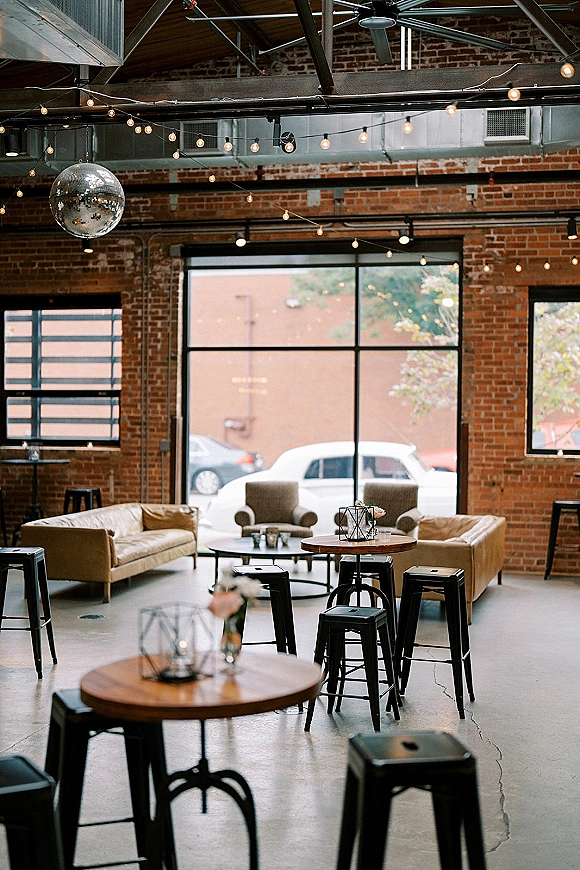 Wedding lounge area with reception lounge seating, sofas and armchairs around cocktail tables under string lights and a disco ball in a brick loft space