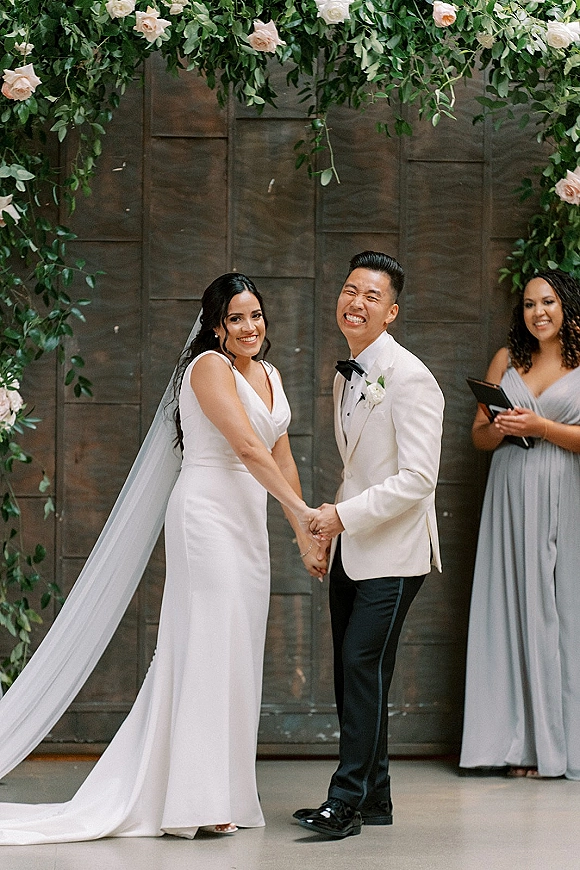Ceremony moment as bride and groom holding hands and smiling under a rose and greenery arch, long veil trailing against wood-paneled wall