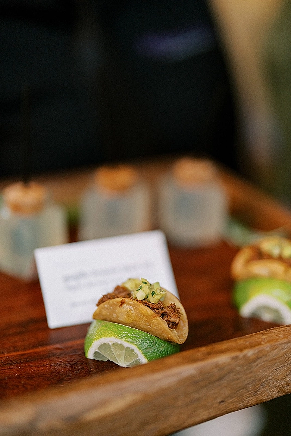 Wedding reception food of mini taco wedding appetizer with lime wedge on a wooden serving board beside place card and votive candles, guests blurred