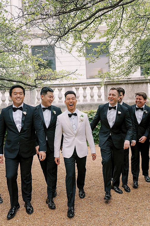Groomsmen portrait of groom in a white dinner jacket with friends in black tuxedos, laughing on a gravel path by an estate facade