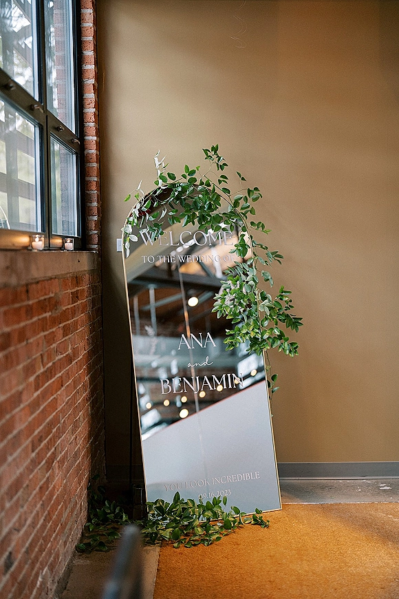 Wedding welcome sign on an arched mirror with ivy garland and candles, leaning against a brick wall by an industrial window