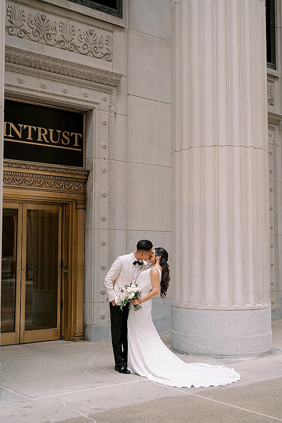Wedding kiss portrait of bride and groom kissing, bride holding a white bouquet before an ornate stone doorway with brass revolving door