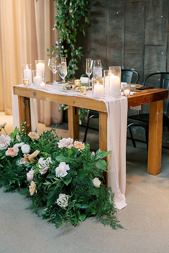 Reception tablescape with head table decor on a wood dining table, pillar candles in glass, champagne flutes, and lush floral greenery runner before drapery curtains