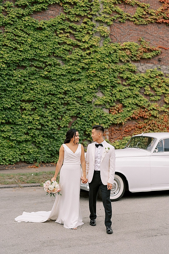 Couple portrait of bride and groom holding hands beside a vintage car, her bouquet and dress train against an ivy-covered brick wall