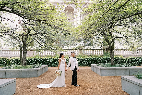 Couple portrait of bride and groom holding hands, bride with bouquet and long dress train beside an ivy wall and stone balustrade in a garden