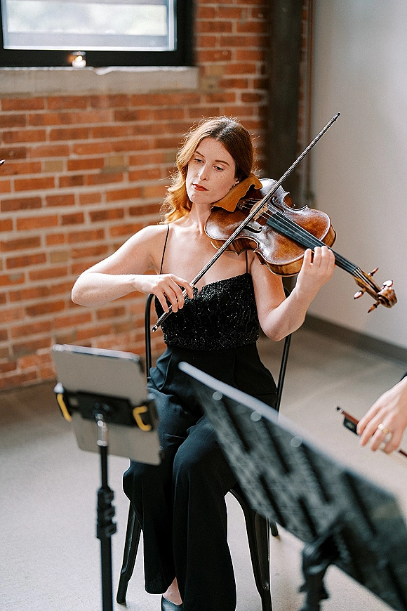 Wedding violinist playing with bow and sheet music on a stand, seated in a black evening dress against a brick wall indoors