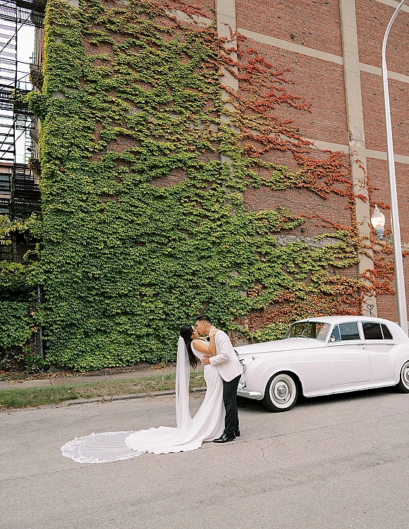 Wedding kiss portrait of bride and groom kissing beside a white classic car, her long veil and lace train flowing by an ivy brick wall