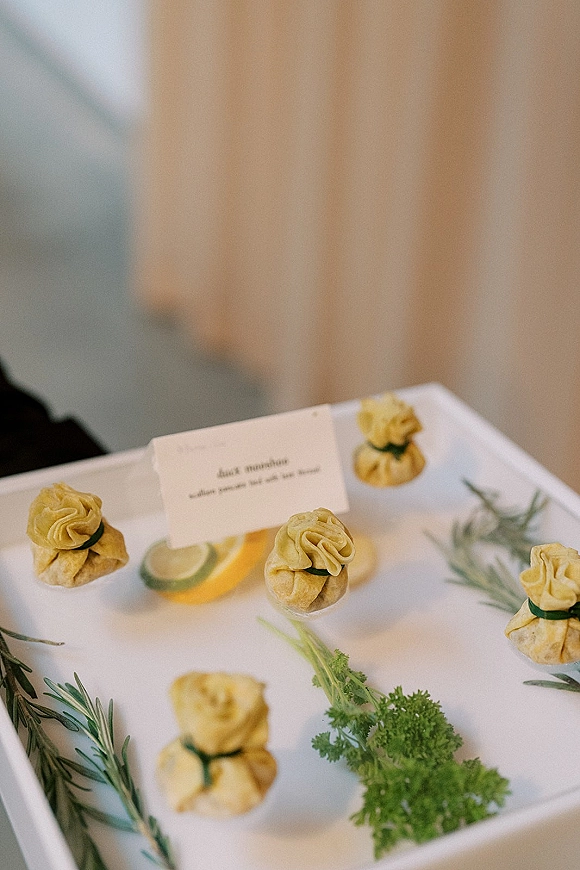 Wedding appetizer platter of dumpling bites on a white serving tray with a place card, lemon slices, and rosemary garnish in soft indoor light