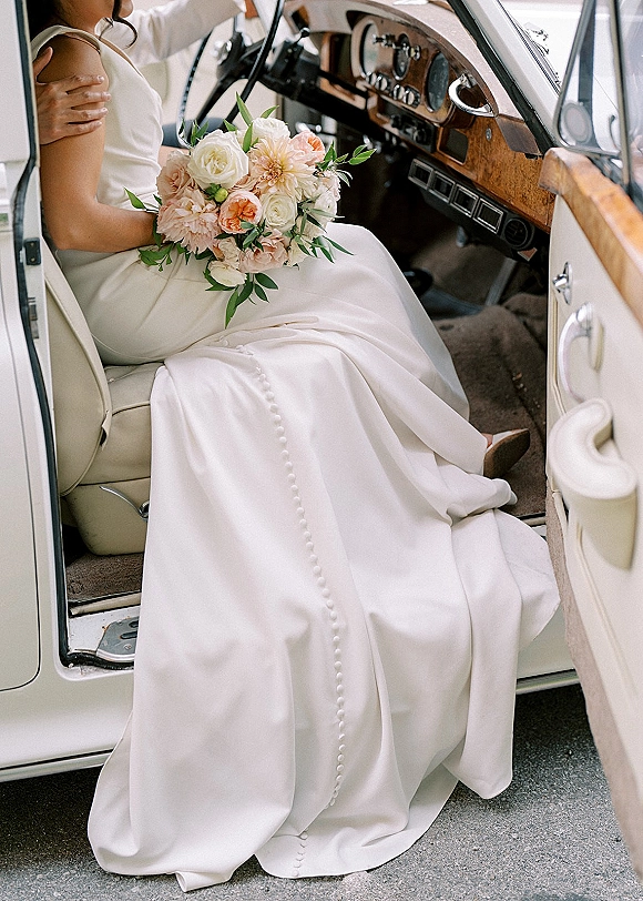 Bride in car holding a wedding car photo bouquet, showing satin dress buttons and ring against leather seats and wood dashboard