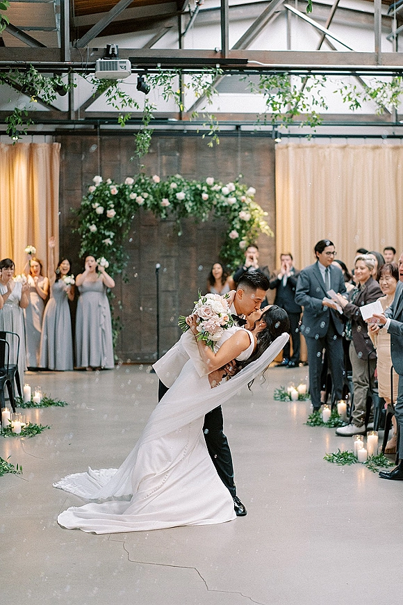 Wedding kiss as the groom dips the bride holding her bouquet, veil flowing beneath a floral arch with candles and cheering guests indoors