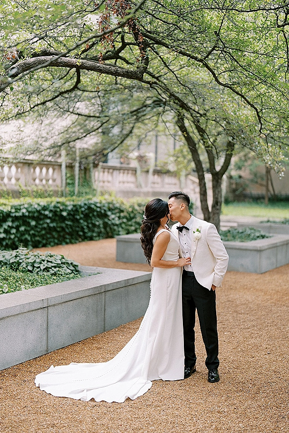 Wedding kiss portrait of bride and groom kissing on a garden walkway, her long train trailing as he wears a white dinner jacket under trees