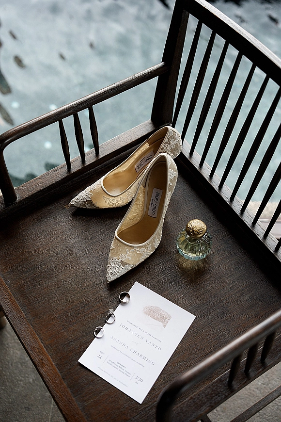 Wedding flat lay with lace bridal heels, invitation suite, rings, and perfume bottle arranged on a stone floor with a wooden chair accent