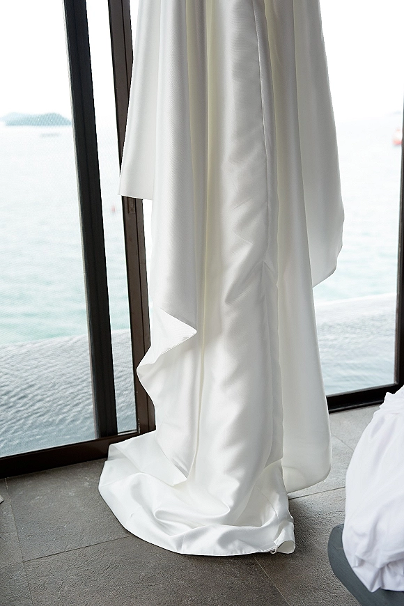 Wedding dress in satin hangs by a floor-to-ceiling window, its long train draped in natural light with an ocean view beyond