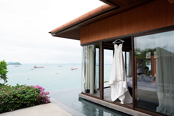 Wedding dress on hanger framed by curtains in sliding glass doors, with a strapless gown overlooking ocean, boats, and villa patio