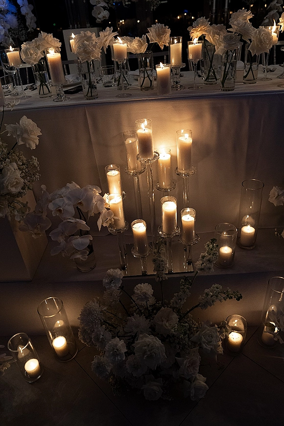 Wedding candle decor with cylinder vase candles and pillar candles beside white orchids on mirrored risers at a dimly lit head table