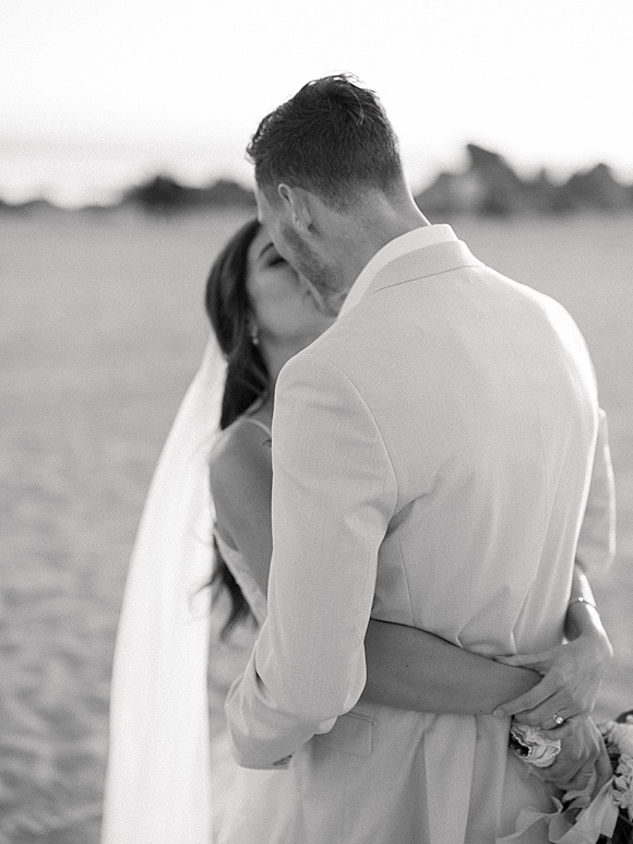 Wedding kiss portrait of newlyweds kissing as the bride’s veil blows in the wind, bouquet in hand, with ocean horizon behind