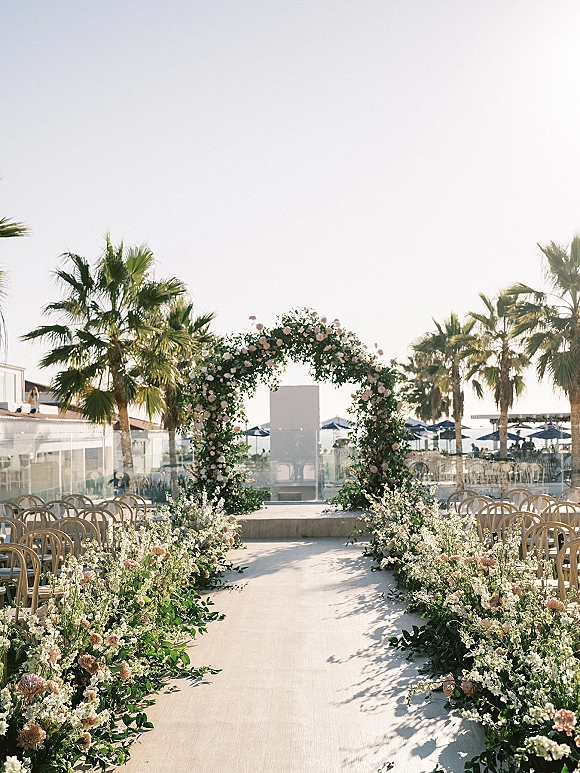 Ceremony aisle decor with a round floral arch and blush-and-white arrangements lining wooden chairs on a palm-lined outdoor terrace